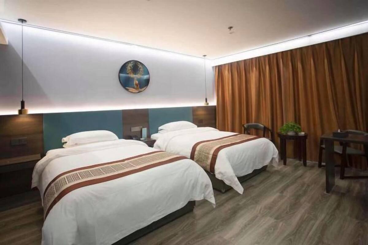 Habitación de hotel con dos camas, un reloj de pared redondo, un cabecero de madera, una mesita con una planta y una cortina.