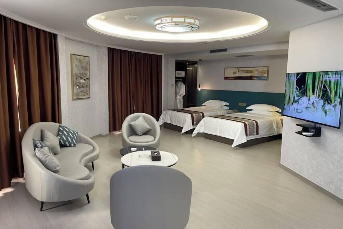 A modern hotel room with a curved sofa, a round coffee table, a bed with a white headboard, a television on the wall, and a painting above the bed.