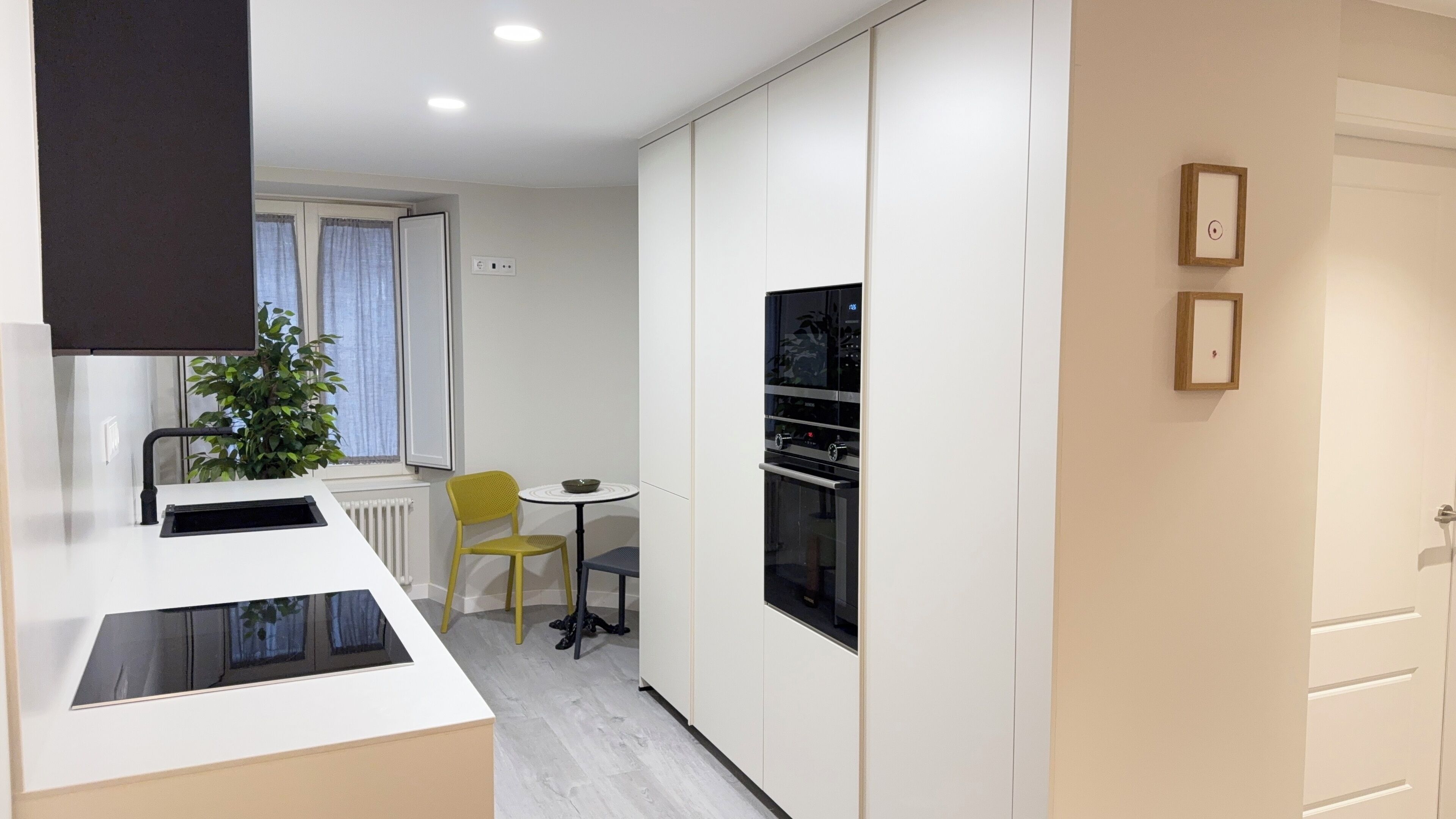 Una cocina moderna con horno empotrado, un pequeño comedor con mesa y sillas, y una planta en maceta.