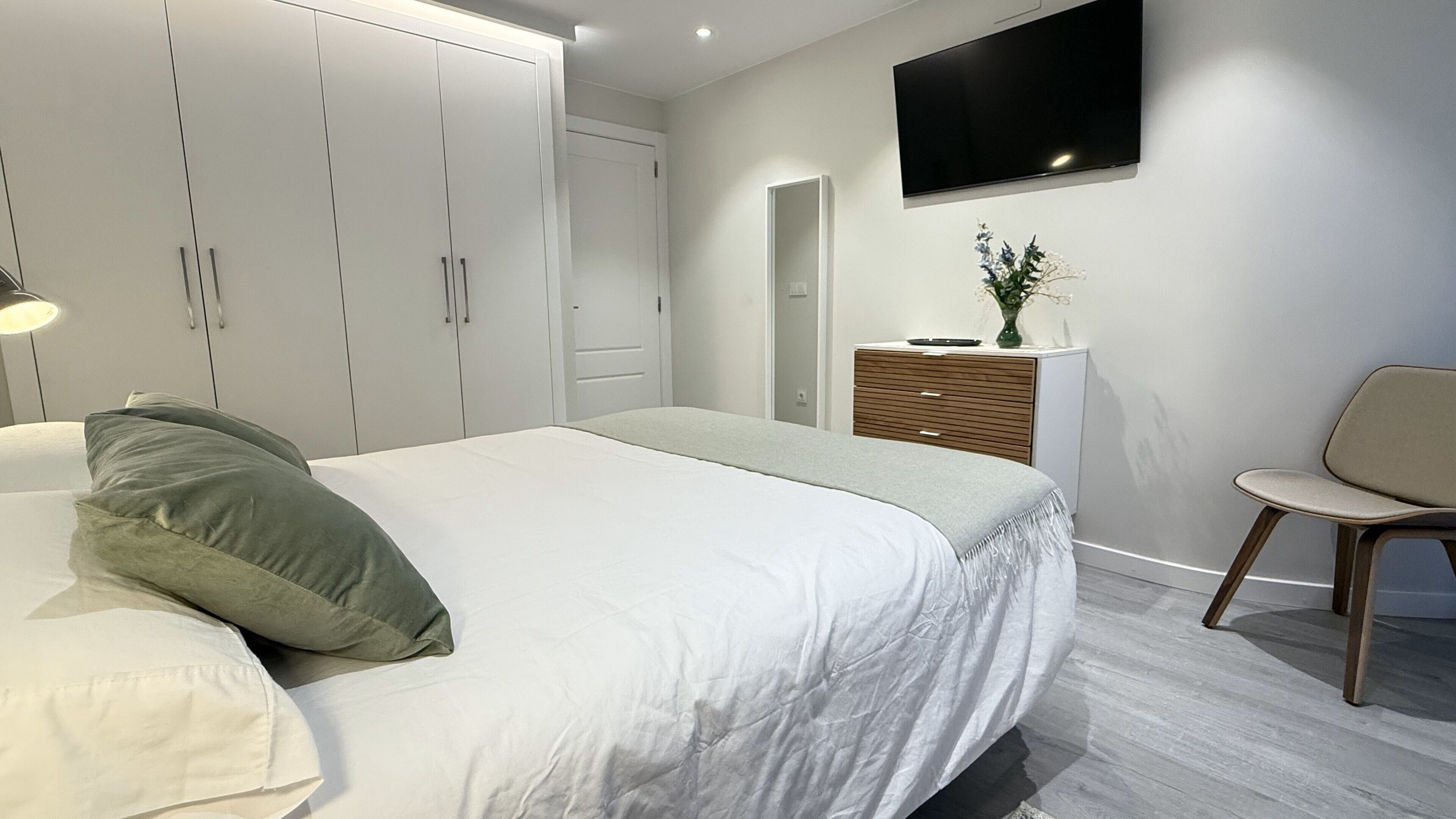 Un dormitorio moderno con una cama, un aparador, una silla y un televisor en la pared.