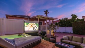 Un patio al aire libre con un sofá exterior con calefacción, una piscina pequeña y una pantalla de televisión mostrando una película.