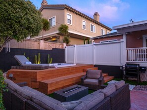 A backyard with a hot tub, wooden deck, outdoor seating, and a white fence.