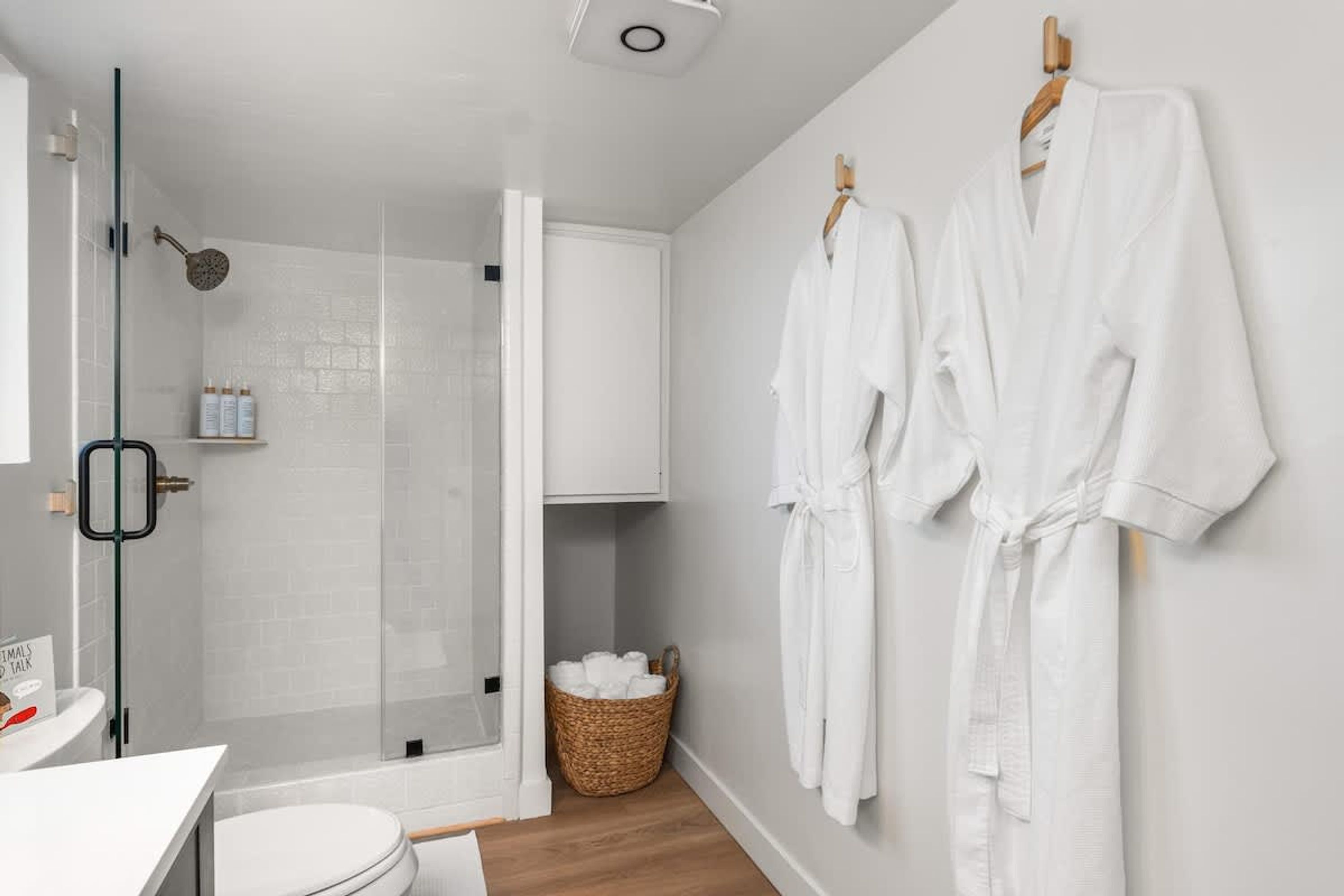 A modern bathroom with a glass shower enclosure, a white sink, and two white robes hanging on a wooden rack.