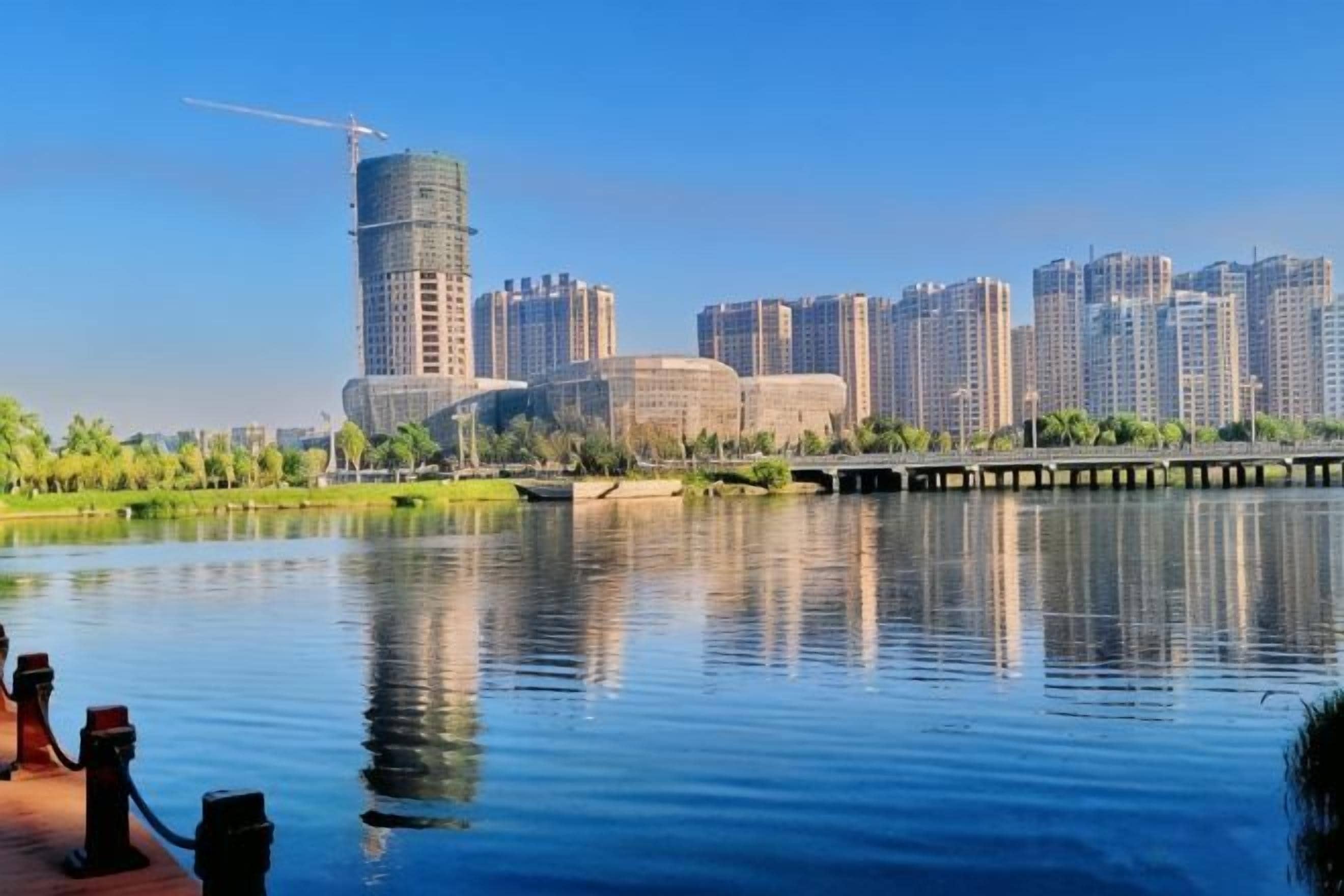 A waterfront cityscape with high-rise buildings, a calm river, and a bridge.