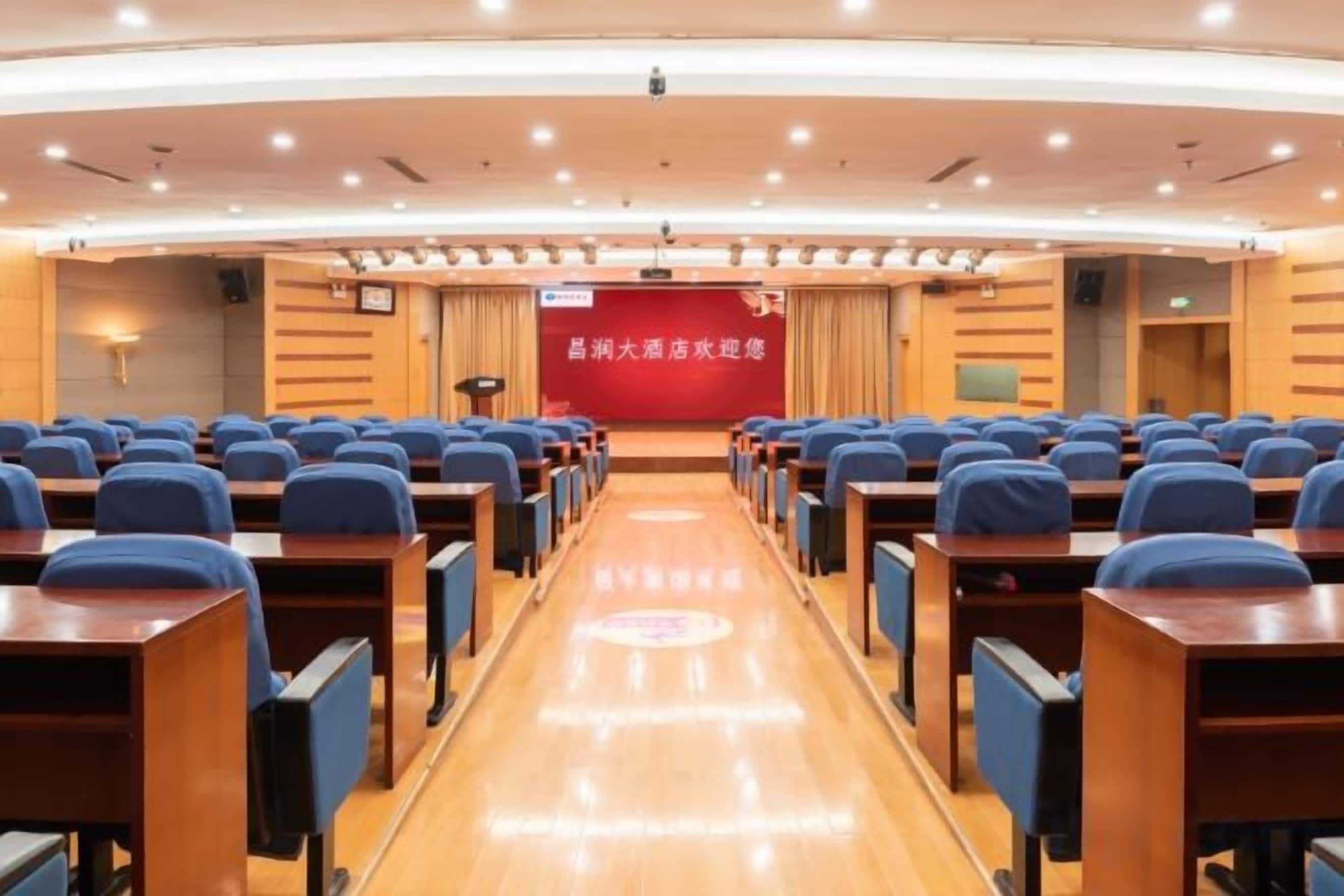 Une salle de conférence avec des rangées de tables et de chaises, une scène dotée d’un fond rouge et un pupitre.