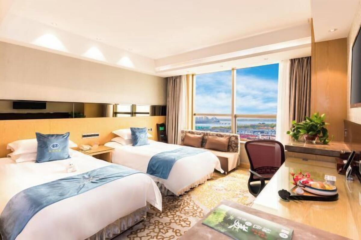 A hotel room with two beds, a desk, a chair, a TV, and a large window with a city view.