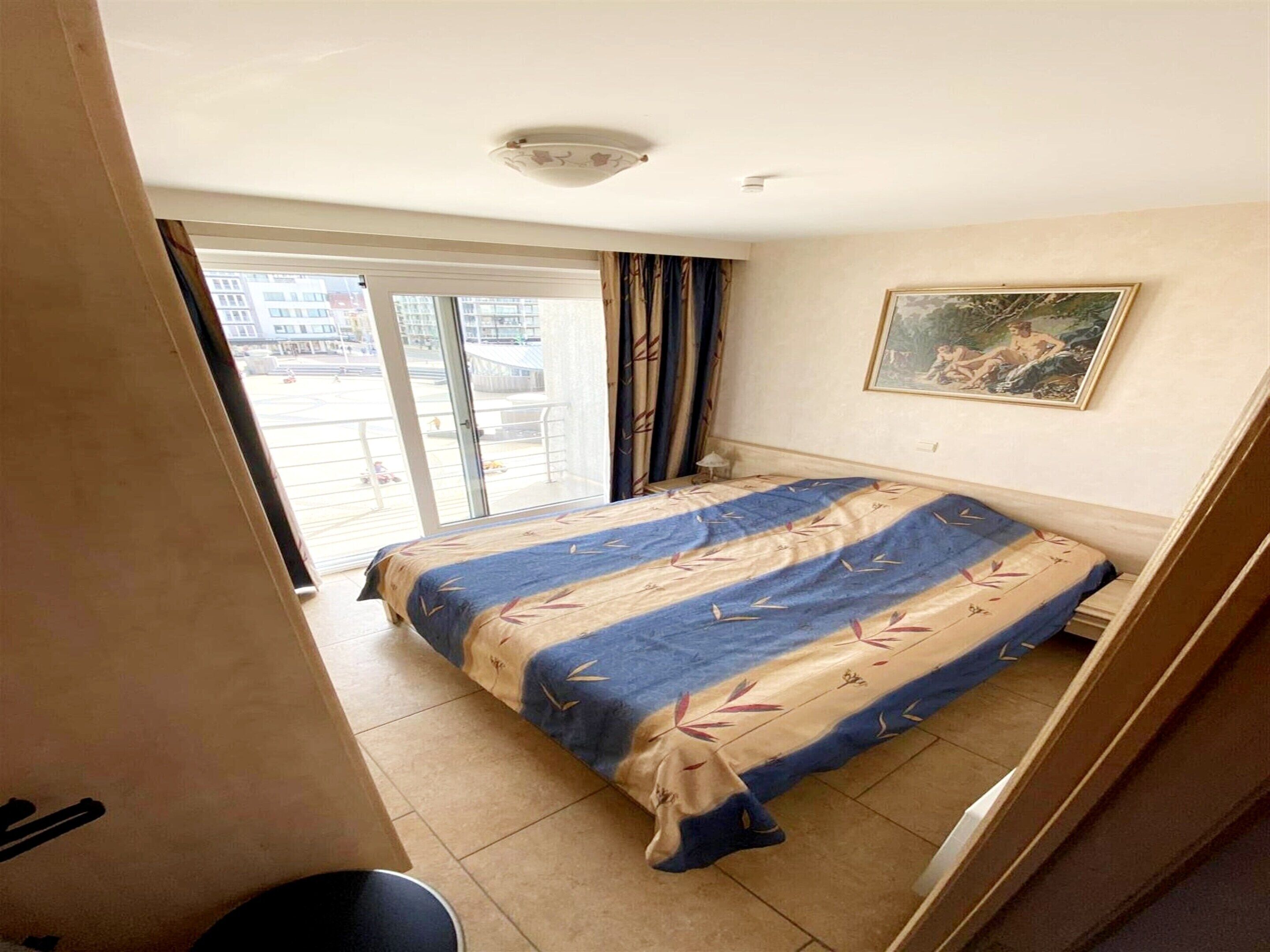 A hotel room with a bed, a painting on the wall, and a view of the outside balcony.