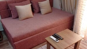 A red sofa with beige cushions, a small wooden table, and a striped rug in a room with a plain wall and a curtain. - Florenze Khamsin Hurghada- - Room K203 (Hurghada)