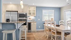 A kitchen with white cabinets, a stainless steel refrigerator, a microwave, and a built-in oven. There is a dining area with a table and chairs, and a window with blinds. - Always Sunday-Pet Friendly Home in Atlantic Beach, NC (Atlantic Beach)