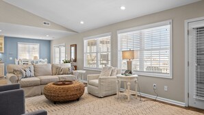 A spacious living room with a beige sofa, a grey armchair, and a woven ottoman. There are large windows with blinds, a lamp on a side table, and a small round table with decorative items. - Always Sunday-Pet Friendly Home in Atlantic Beach, NC (Atlantic Beach)