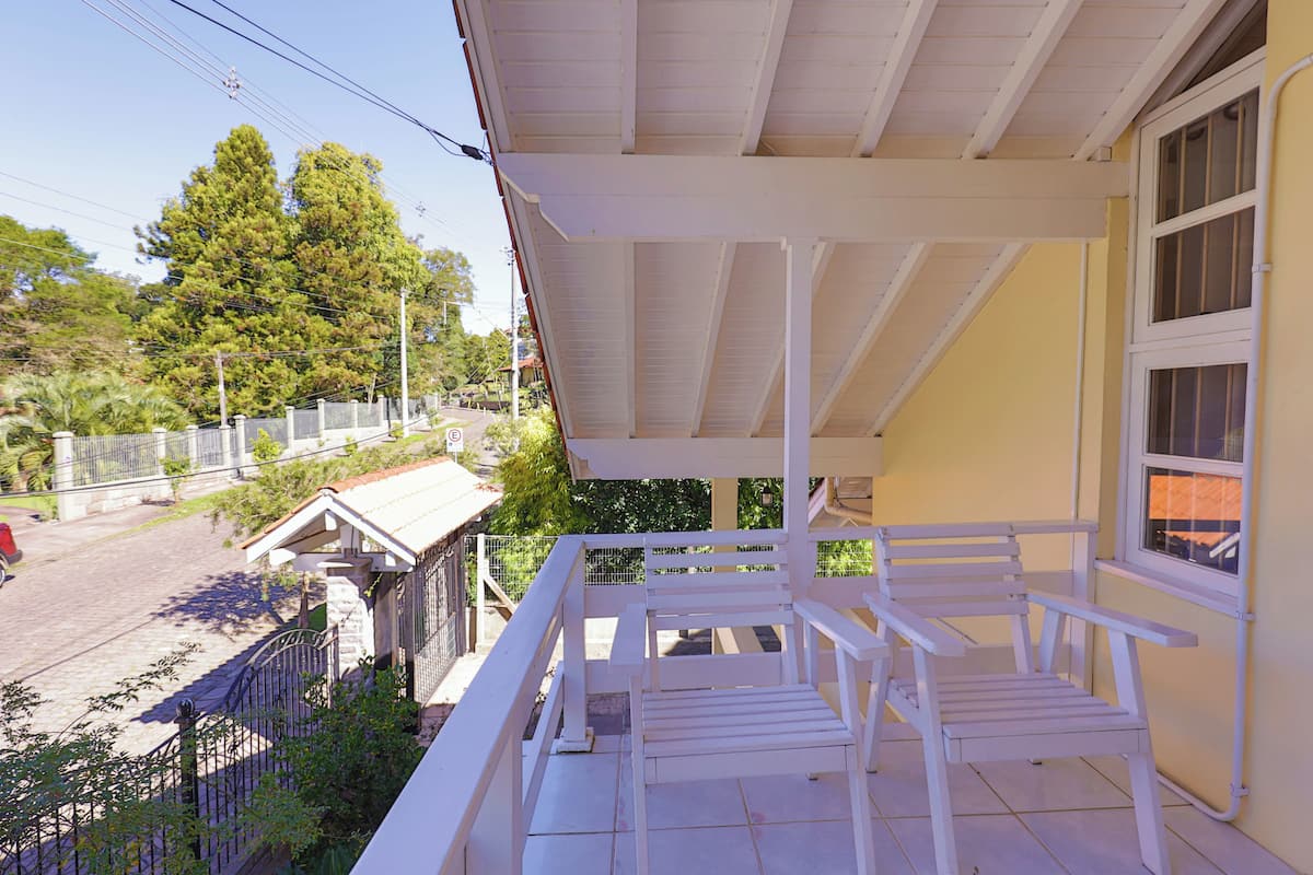 A covered balcony with white railings and a bench, overlooking a street with trees and a fence.