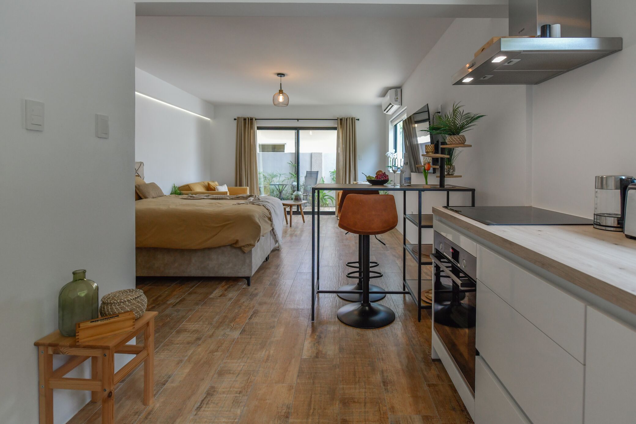 A modern hotel room with a bed, a small dining table, a kitchen area, and a view of the outdoors.