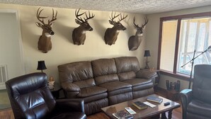 Ein Wohnzimmer mit einem braunen Ledersofa, zwei Ledersesseln, einem Couchtisch mit Büchern und vier aufgehängten Hirschköpfen an der Wand.