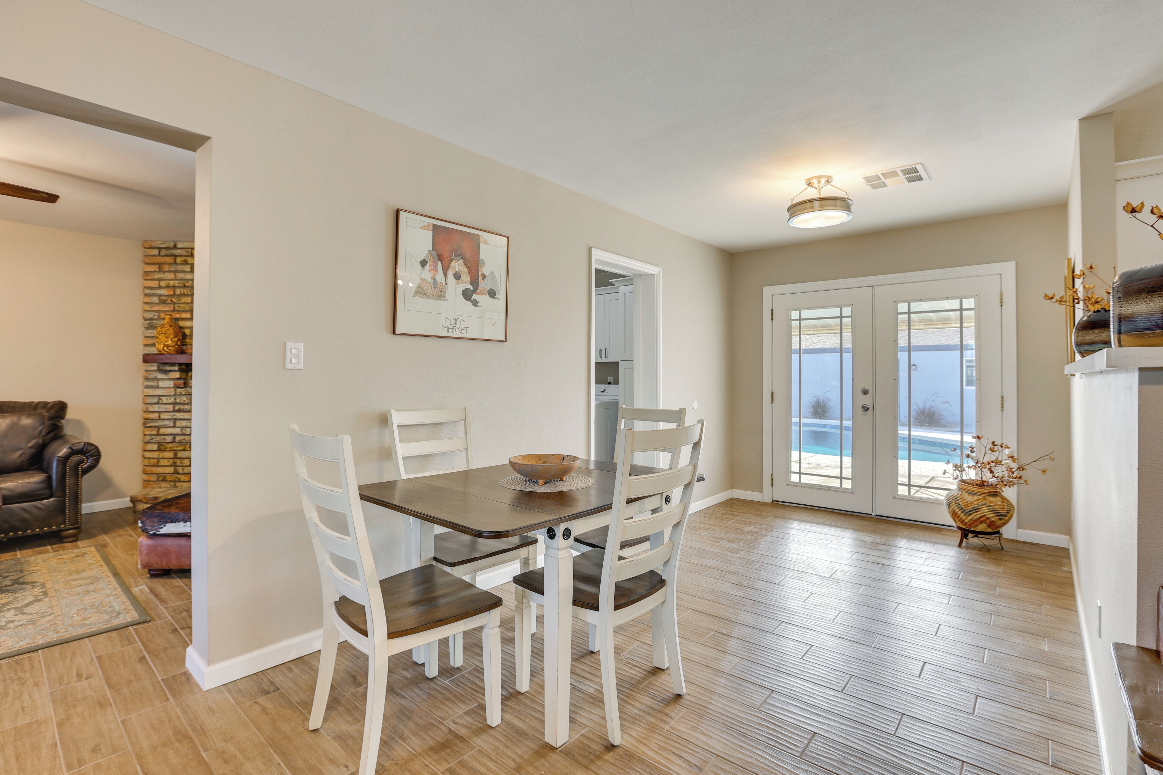 Un espace repas avec une table en bois et des chaises blanches, un mur décoratif en briques et une vue sur la piscine à travers des portes vitrées.