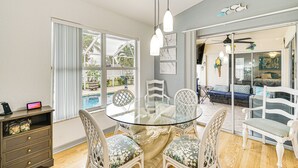 A dining area with a glass table surrounded by wicker chairs, a dark wooden cabinet, and a view of a swimming pool through large windows.