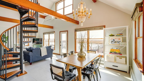 A spacious living area with a wooden staircase, a chandelier, a dining table with chairs, and a bookshelf. - 2 Mi to Stone Harbor Beach: Lovely Bayfront Home (Cape May Court House)