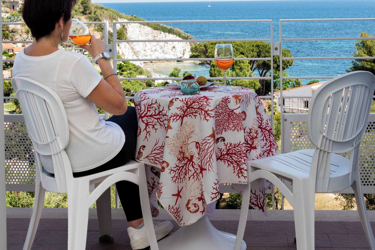 Eine Person sitzt an einem Tisch mit Blick aufs Meer, eine Tischdecke mit rotem Muster und ein Glas mit Getränk.