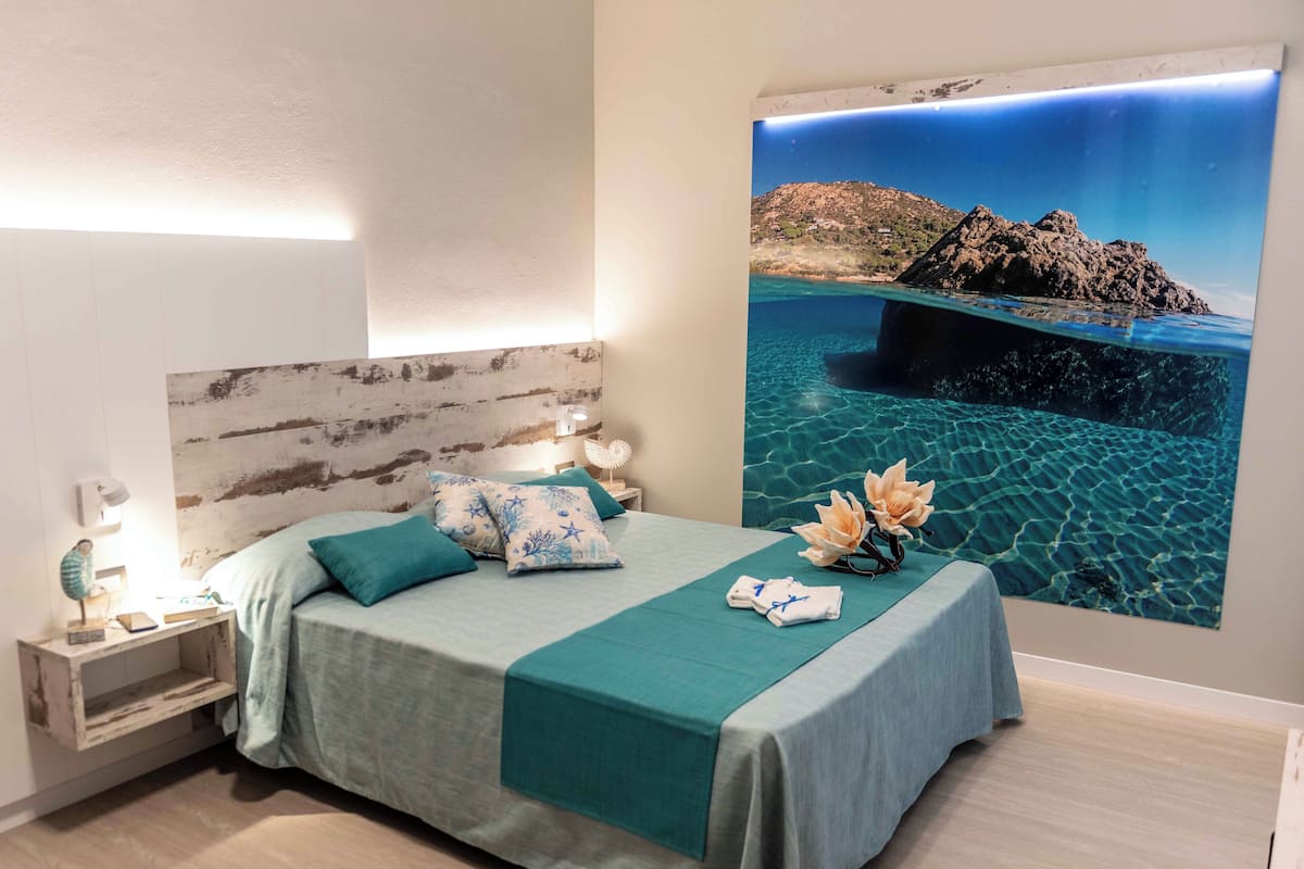 Ein Schlafzimmer mit einem Bett, Kissen, einem Nachttisch, einer Wandlampe und einem großen Poster mit einer Küstenansicht.