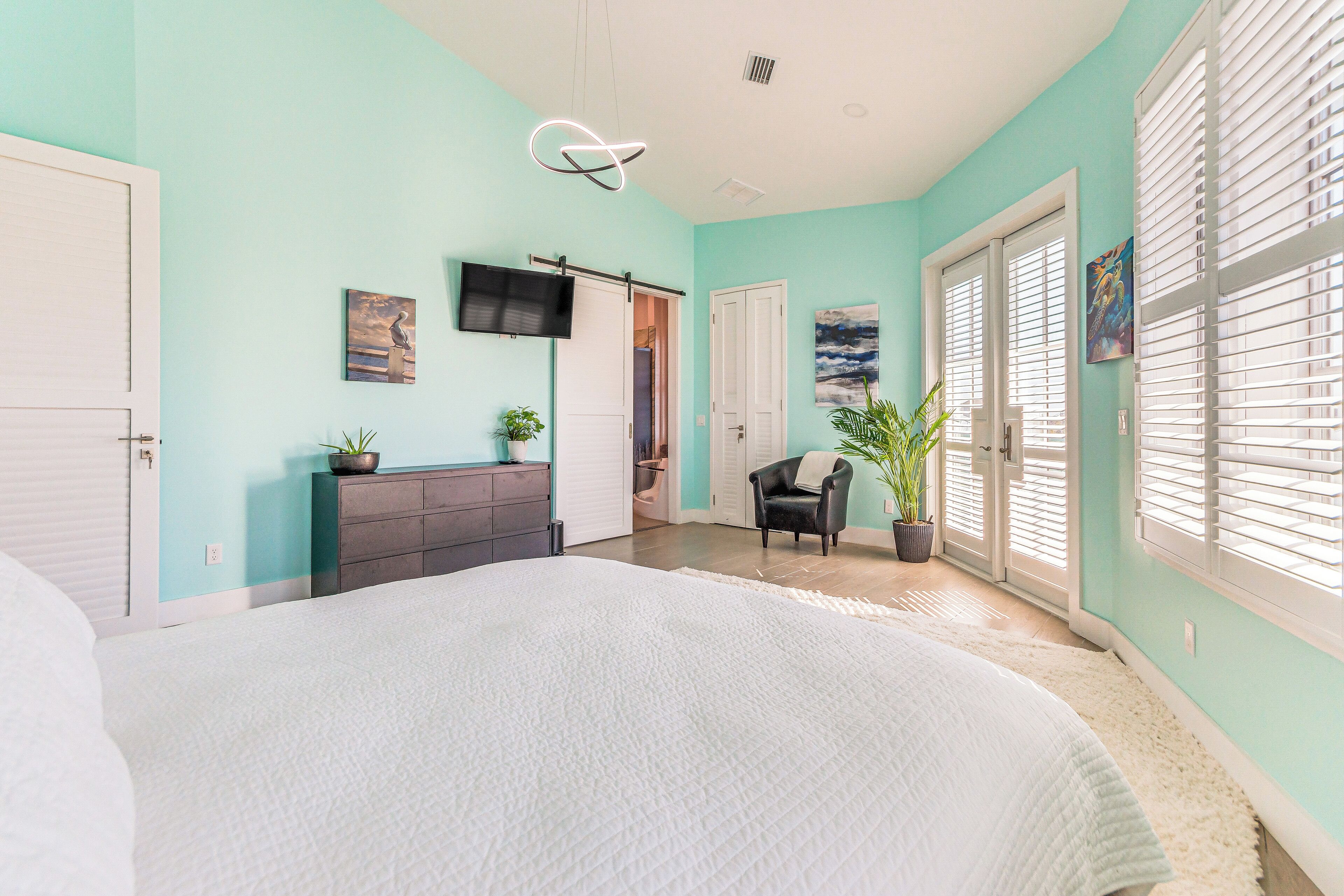 A bedroom with a large bed, a dresser, a TV mounted on the wall, and a door leading to another room.