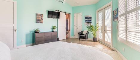 A bedroom with a large bed, a dresser, a TV mounted on the wall, and a door leading to another room.