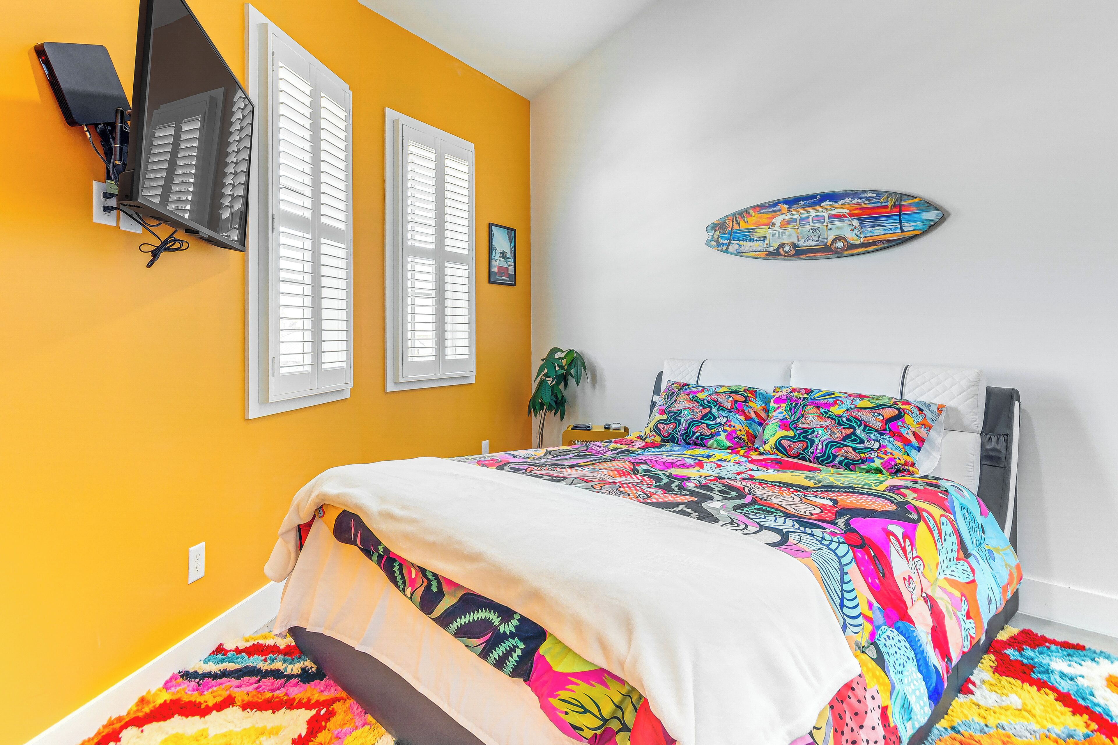 Une chambre à coucher avec un lit, un téléviseur fixé au mur, une fenêtre avec des stores et une décoration murale colorée.