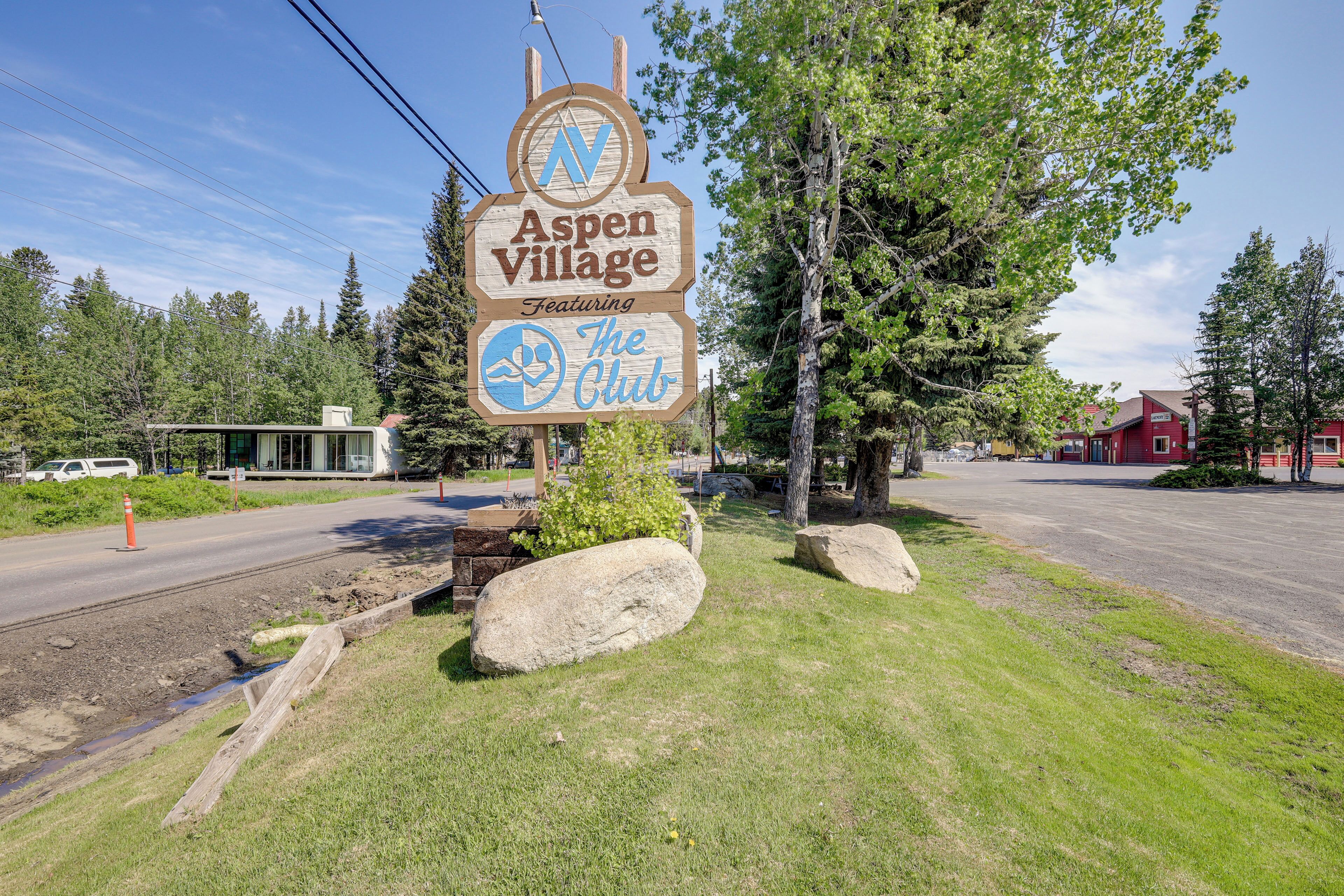 A sign for Aspen Village featuring The Club, surrounded by trees and a red building in the background.
