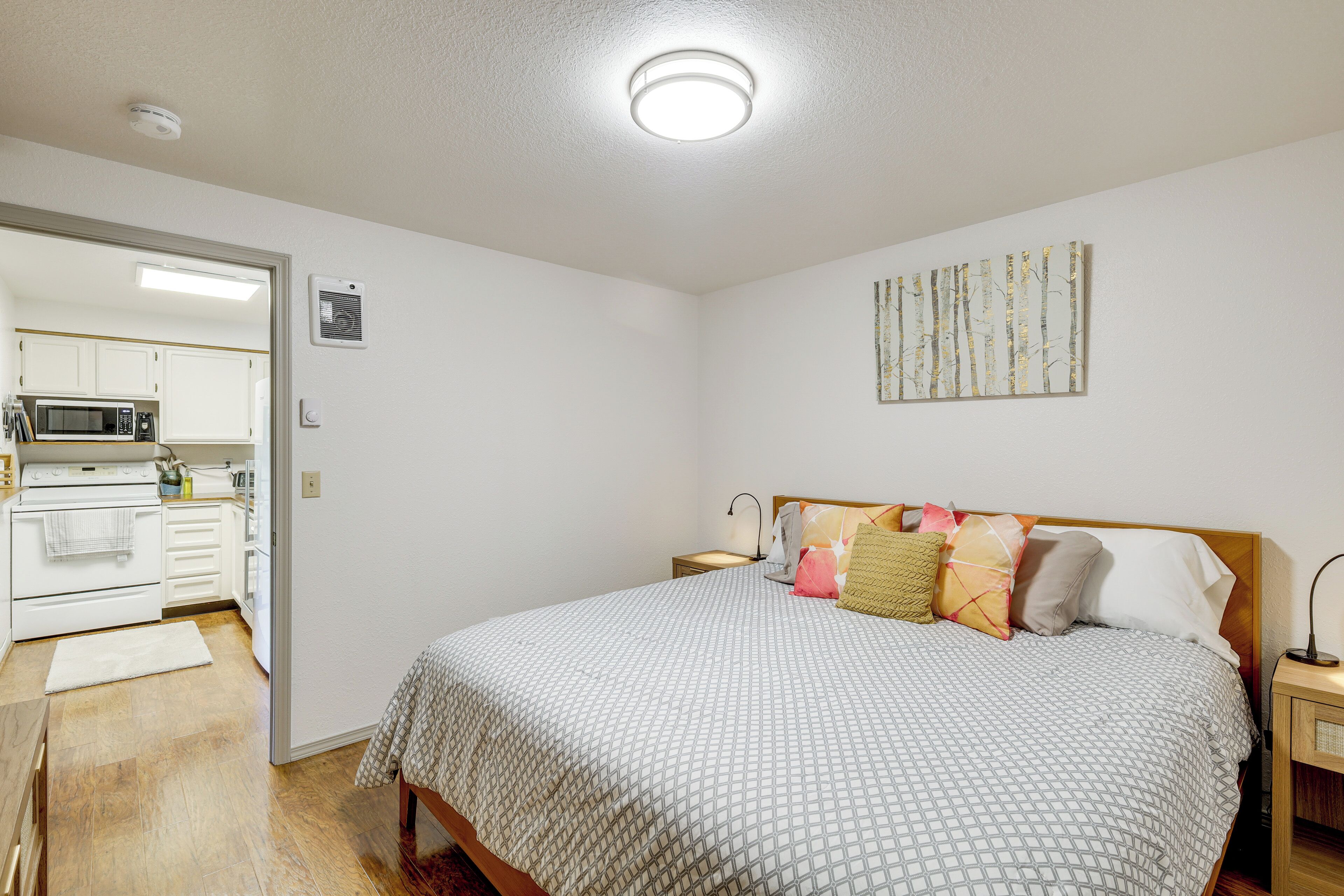 Quarto com cama, mesa de cabeceira, luminária e vista para uma cozinha.