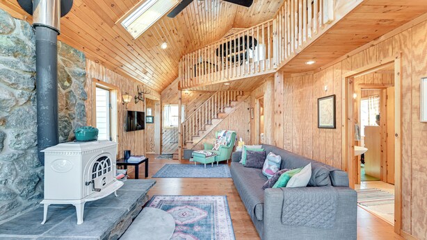 Une cabane en bois spacieuse, dotée d’un mur en pierre, d’un poêle blanc, d’un canapé gris et d’un escalier menant à une mezzanine.