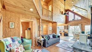 L’intérieur d’un chalet en bois comprend une chambre à coucher située en mezzanine, un salon avec un canapé et un fauteuil, ainsi qu’un coin repas avec un poêle.