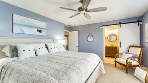 Un dormitorio con una cama grande, un ventilador de techo, un cuadro de un barco en la pared y una silla con cojín.