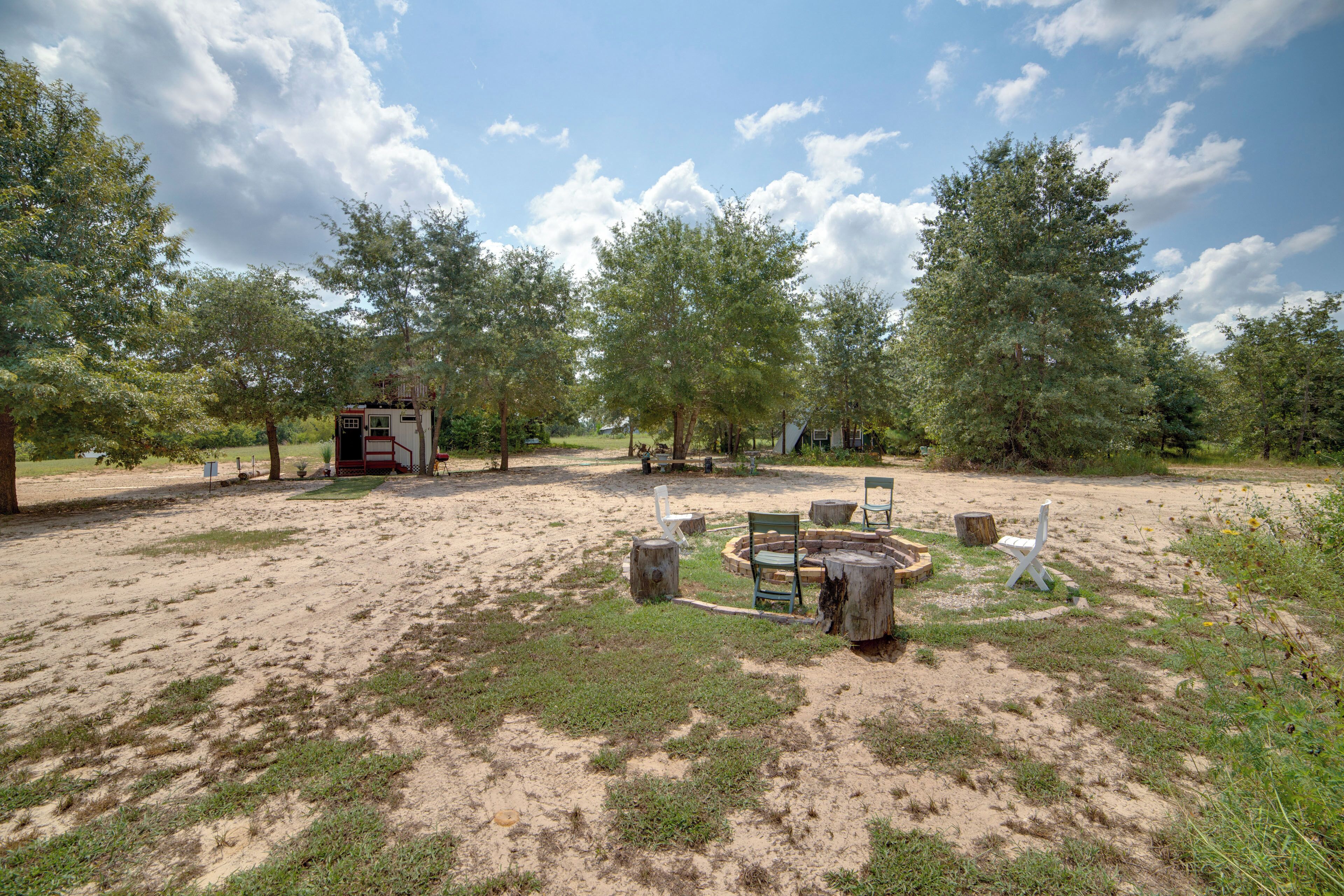 A campsite with a circular seating area surrounded by trees, a small cabin, and a red vehicle in the background.