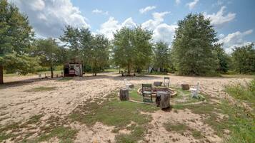 A campsite with a circular seating area surrounded by trees, a small cabin, and a red vehicle in the background.