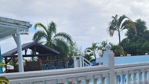 A white balustrade with a pool in the background, palm trees, and a covered area with stairs. - L'ECRIN BLEU SEA VIEW DIAMOND ROCK (LE DIAMANT)