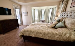 Ein Schlafzimmer mit einem großen Bett, einem Schminktisch, einem Fernseher und einem Fenster mit Vorhängen.