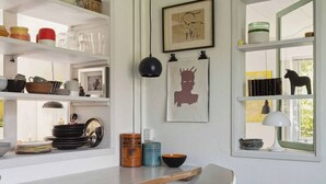 A kitchen with white cabinets, a wooden bar counter, and various kitchenware. There are framed artworks on the wall and a window with a view of greenery. - Furnished Home by Sea & Fjords - Perfect for Remote Work Coastal Artist's Haven (Hundested)