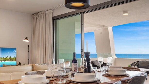 Un coin repas avec une table mise pour le repas, une grande fenêtre donnant sur l’océan et un canapé avec vue sur une plage tropicale.