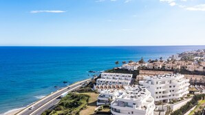 Une zone résidentielle côtière avec des bâtiments modernes blancs, une route parallèle à la plage et une mer d’un bleu limpide.