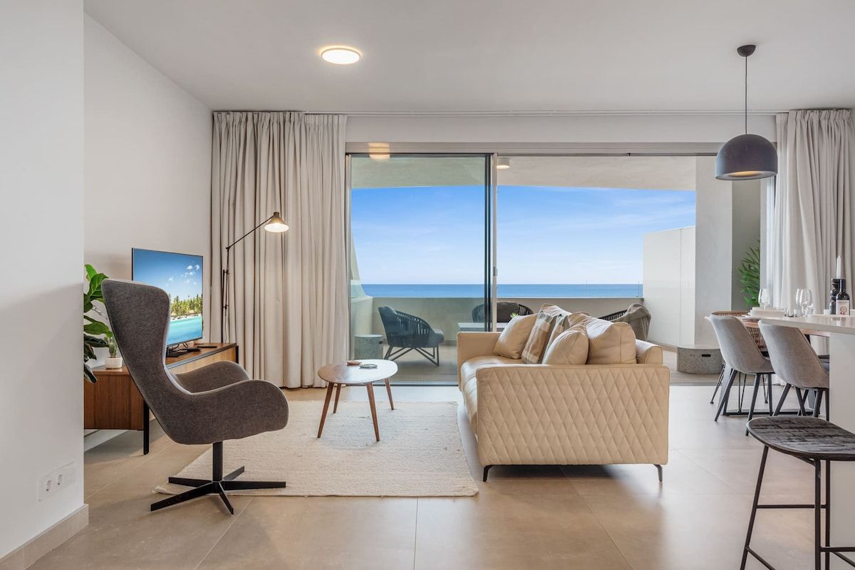 Un salon moderne comprenant un canapé, un fauteuil et un coin repas, avec une grande porte-fenêtre coulissante donnant sur un balcon avec vue.