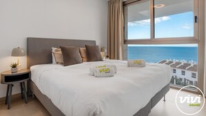 Une chambre d’hôtel avec un grand lit, une table de chevet avec une lampe et une vue sur la mer par une grande fenêtre.