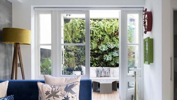 A room with a blue sofa, a floor lamp, and a view of a lush garden through large windows.