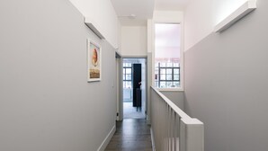 Un pasillo estrecho con barandilla blanca, una puerta al final y una ventana a la derecha.