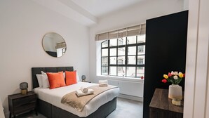 Un dormitorio moderno con una cama grande, una mesita de noche, una ventana con vistas a edificios y un espejo en la pared.