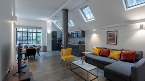 Una sala moderna con un sofá gris, un sillón amarillo y una mesa de centro. Hay tragaluces, una zona de cocina y una mesa de comedor con sillas.