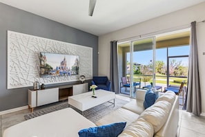 A modern living room with a flat-screen TV, a sofa, a coffee table, and a sliding glass door leading to an outdoor patio. - Kissimmee Retreat: Cozy 2BR Condo near Theme Parks (Kissimmee)