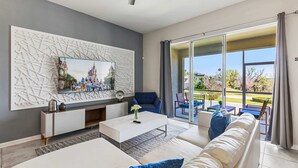Una sala de estar moderna con un televisor de pantalla plana grande montado en una pared texturizada, un sofá blanco con cojines azules, un sillón azul y una puerta de vidrio que da a un patio al aire libre.