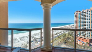 A balcony with a clear view of a beach and ocean, a tall building in the background, and a palm tree-lined street.