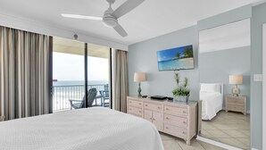 A hotel room with a large bed, a ceiling fan, a balcony with a chair, a painting of a beach, and a dresser with lamps. - Seaside Beach & Racquet 4707 by Liquid Life (Orange Beach)