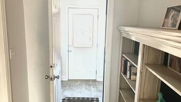 A hallway leading to a door with a doormat, a white wall, and a bookshelf with books and a small figurine.