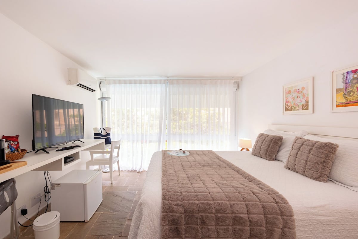Une chambre d’hôtel moderne, équipée d’un lit, d’un bureau avec une télévision et d’une fenêtre avec des rideaux légers.