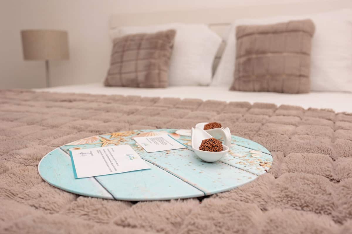 Une table ronde de couleur bleu clair, dotée d’un revêtement rustique, est posée sur un tapis beige moelleux, au milieu d’un lit recouvert de draps blancs et agrémenté de deux coussins marron.